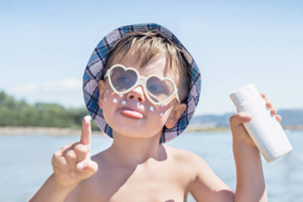 Boy in hat and sunglasses having fun at the beach putting on sunscreen.