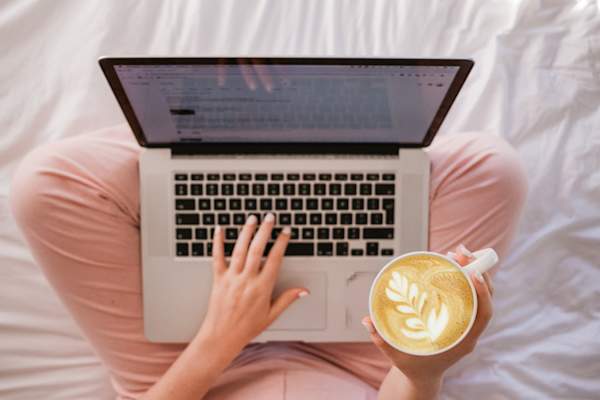 woman sitting on bed, holding latte, looking at laptop