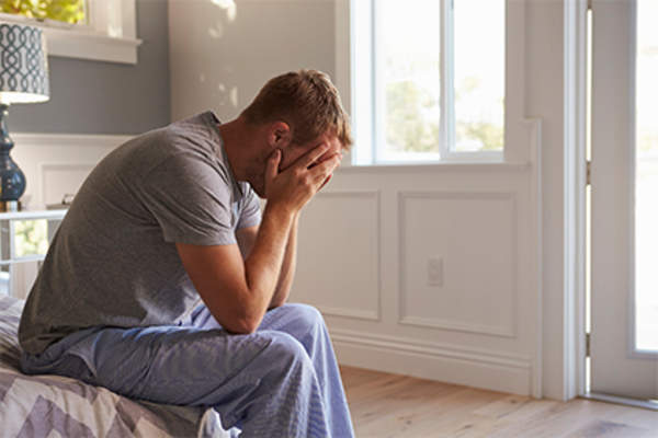 Young man at home in pajamas sitting on bed, worried and tired.