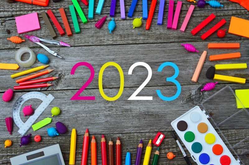 Stół, na nim przybory szkolne, materiały plastyczne, farby, taśmy klejące, kolorowe karteczki. W środku napis 2023