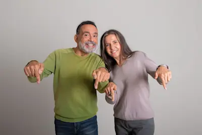 Pareja de adultos mayores sonriente señala hacia abajo con ambas manos