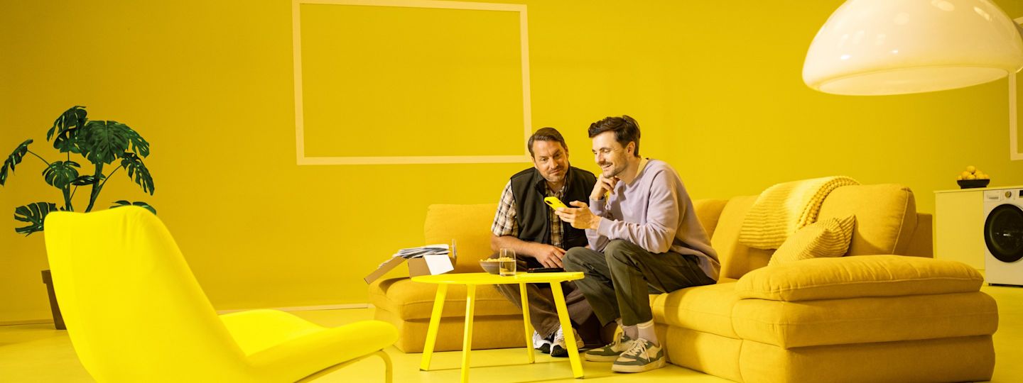 Unser Online Service für dich - ganz ohne Wartschleifen Zwei Männer auf einem gelben Sofa schauen zusammen aufs Smartphone