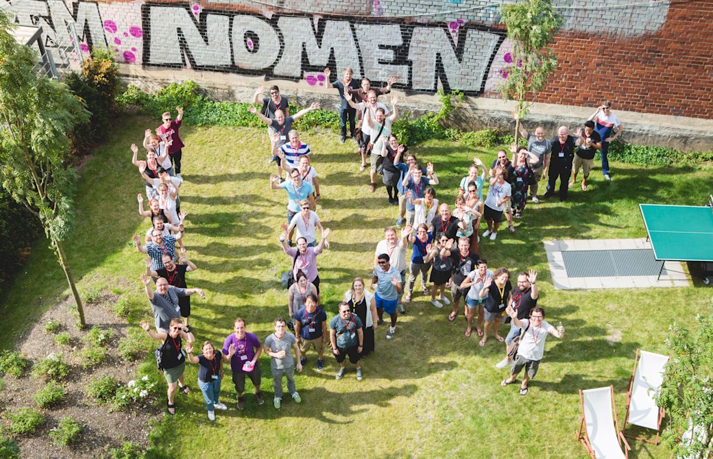 Eine große Gruppe von Menschen steht draußen auf einer Wiese zusammen und bilden die Form der Buchstaben "UX".