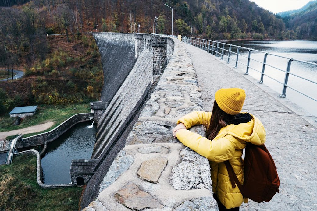 Mit Wasserkraft lässt sich Ökostrom zuverlässig erzeugen Eine Frau mit gelber Jacke und Mütze steht auf einem Staudamm in den Bergen