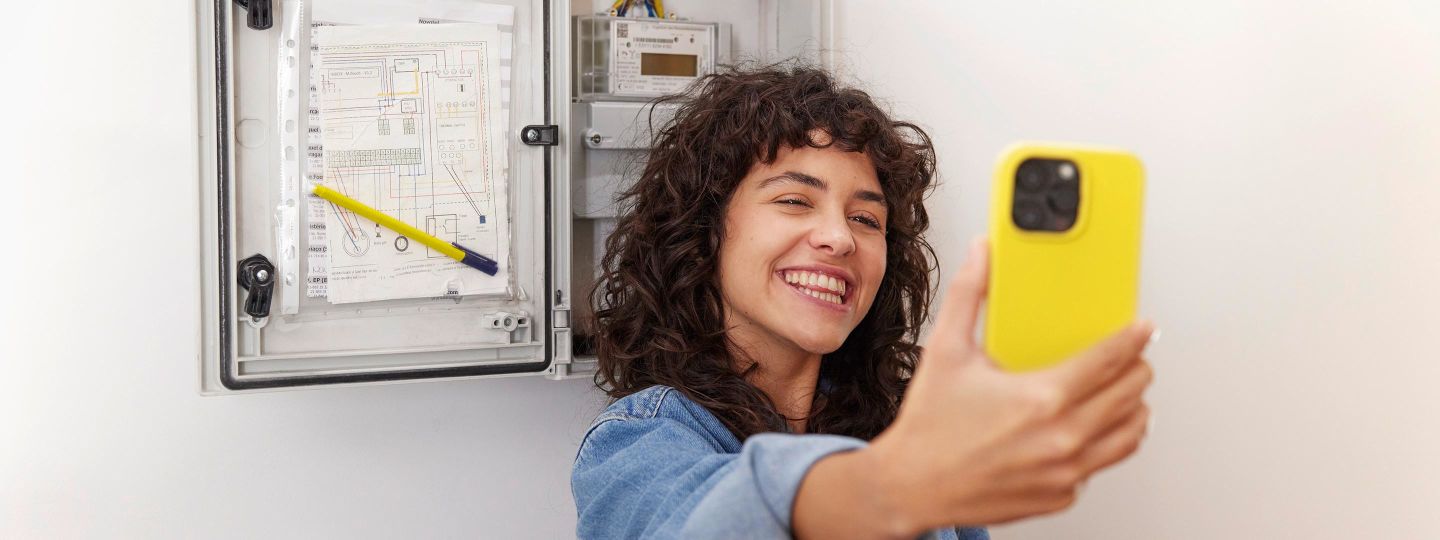 Frau steht mit gelbem Handy vor dem Stromzähler und macht ein Selfie