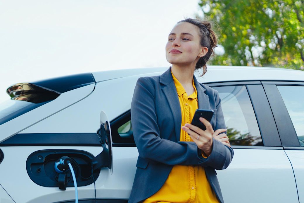E-Auto: Den elektrischen Firmenwagen schnell zuhause laden. Junge Frau mit Smartphone beim Laden ihres Elektroautos