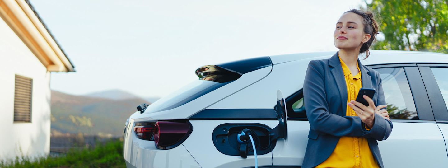 Mit Autostrom von Yello lädst du zuhause und unterwegs ganz entspannt Junge Frau mit Smartphone beim Laden ihres Elektroautos