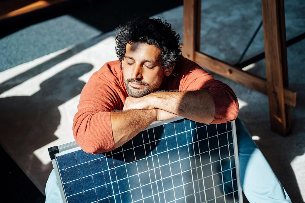 Super praktisch und gut beraten. Das Solarangebot für Miete oder Kauf über Yello  Mann mit orangem Pullover lehnt entspannt auf einem Solarmodul
