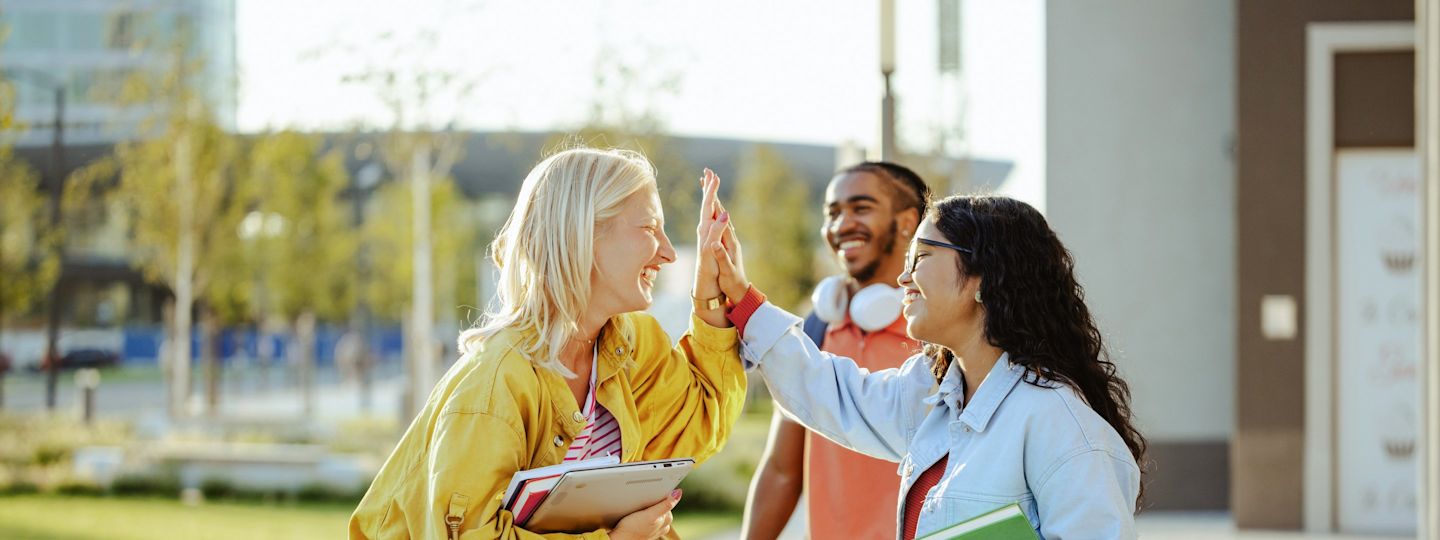 Strom und Gas für Studis günstig bei Yello beziehen Zwei junge Frauen geben sich lachend ein High-Five, ein Freund mit Skateboard begleitet sie im Hintergrund
