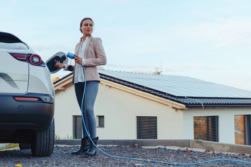 Mit Sonnenstrom lädst du dein E-Auto noch nachhaltiger Eine Frau, gekleidet in Jeans, weißer Bluse und Blazer, steht an einem weißen Elektroauto und hält ein Ladekabel in der Hand