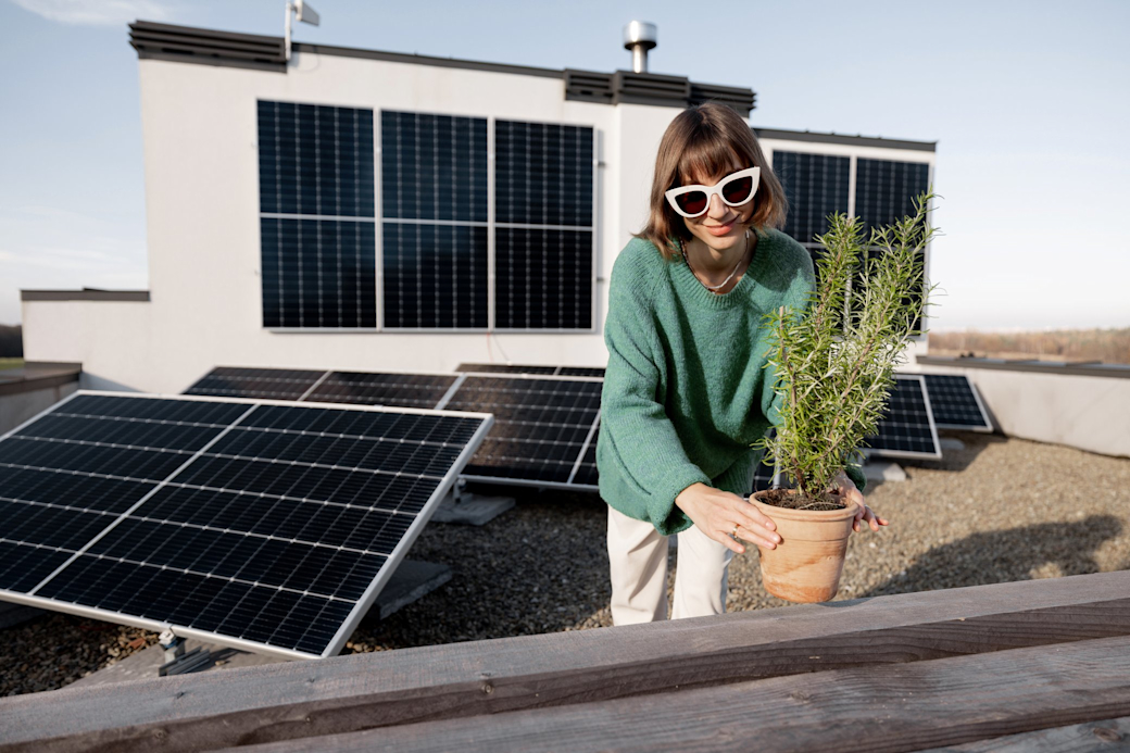 Mit einer eigenen Solaranlage machst du deine eigene grüne Energie Frau mit Sonnenbrille setzt eine Pflanze auf eine Dachterrasse mit Solarmodulen
