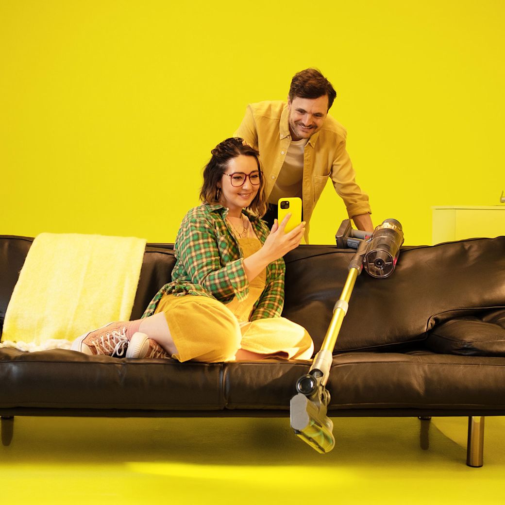 Mit dem Kombitarif Strom und Gas bis du günstiger unterwegs Junge Frau auf Couch zeigt Mann mit Staubsauger ihr Smartphone.