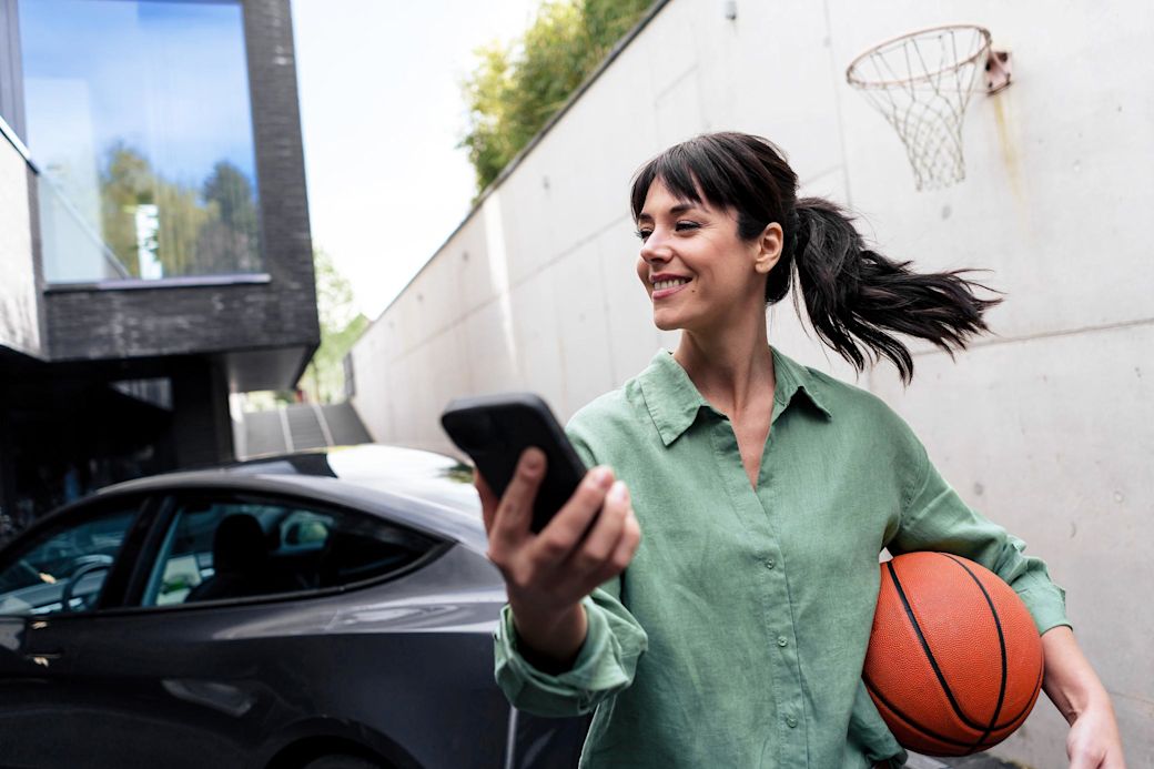 Der dynamische Tarif von Yello bringt mehr Flexibilität in dein Zuhause Frau mit Basketball und Handy vor ihrem E-Auto