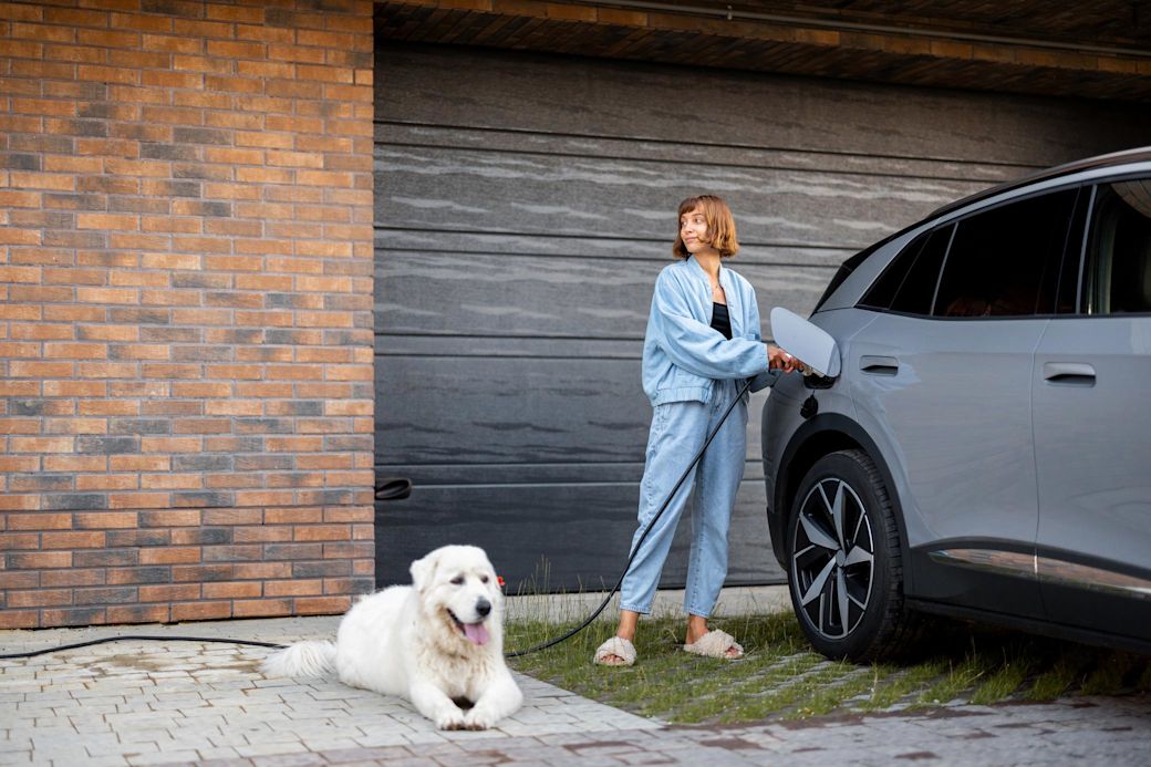 Im Smart Home mit E-Auto und Energiemanagement Strom effizient nutzen Frau lädt ihr Elektroauto zuhause, ein weißer Hund wartet