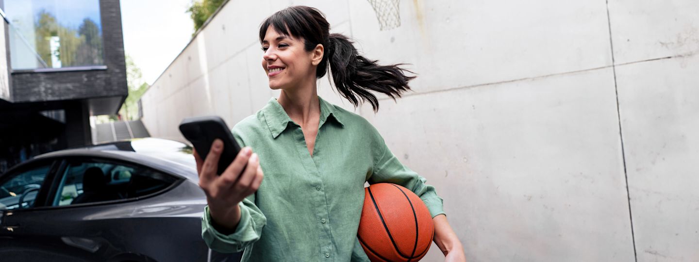 Der dynamische Tarif von Yello bringt mehr Flexibilität in dein Zuhause Frau mit Basketball und Handy vor ihrem E-Auto