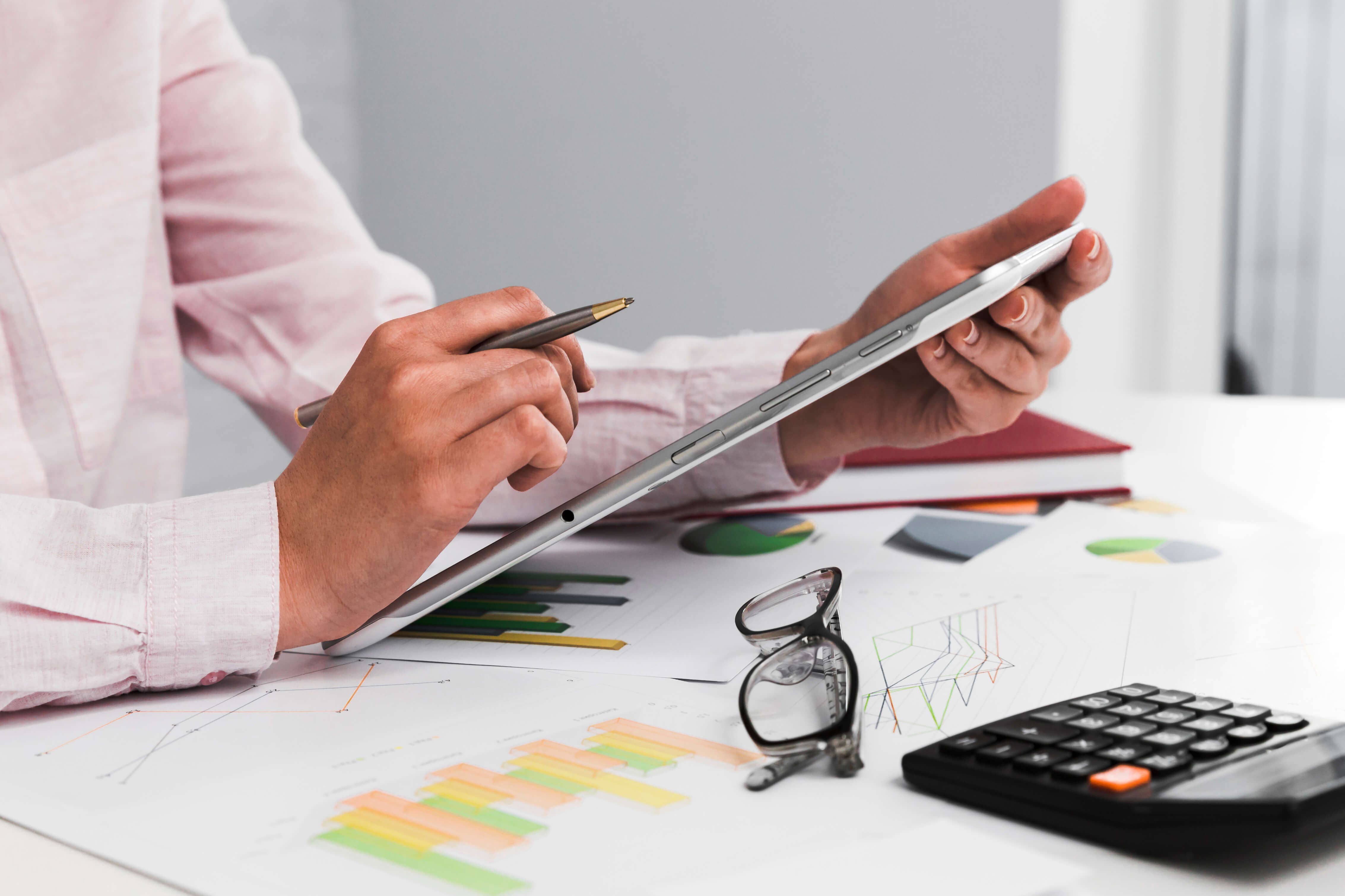 Person reviewing data on a tablet with charts and graphs on printed documents, a calculator and eyeglasses on the desk.