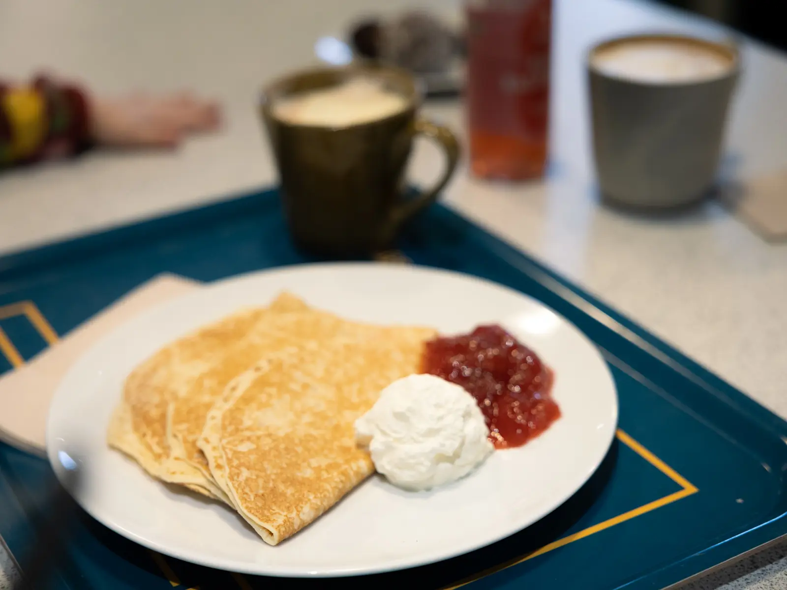 En tallrik med pannkakor, vispad grädde och sylt på. Tallriken ligger på en blå bricka med gula detaljer. Bredvid tallriken står en fylld mugg. I förgrunden kan vi se en rosa flaska, ytterligare en mugg och ett barns ena hand.