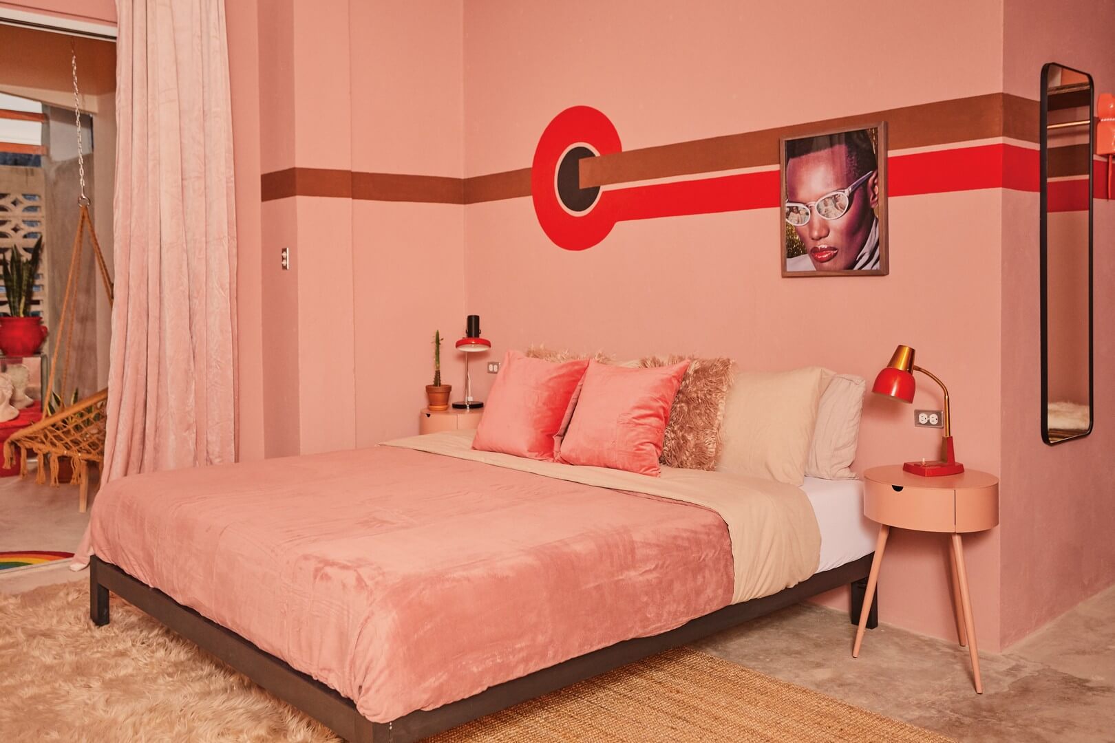 A retro-themed bedroom with pink walls, a pink bedspread, a red accent stripe, and a framed portrait above the bed.