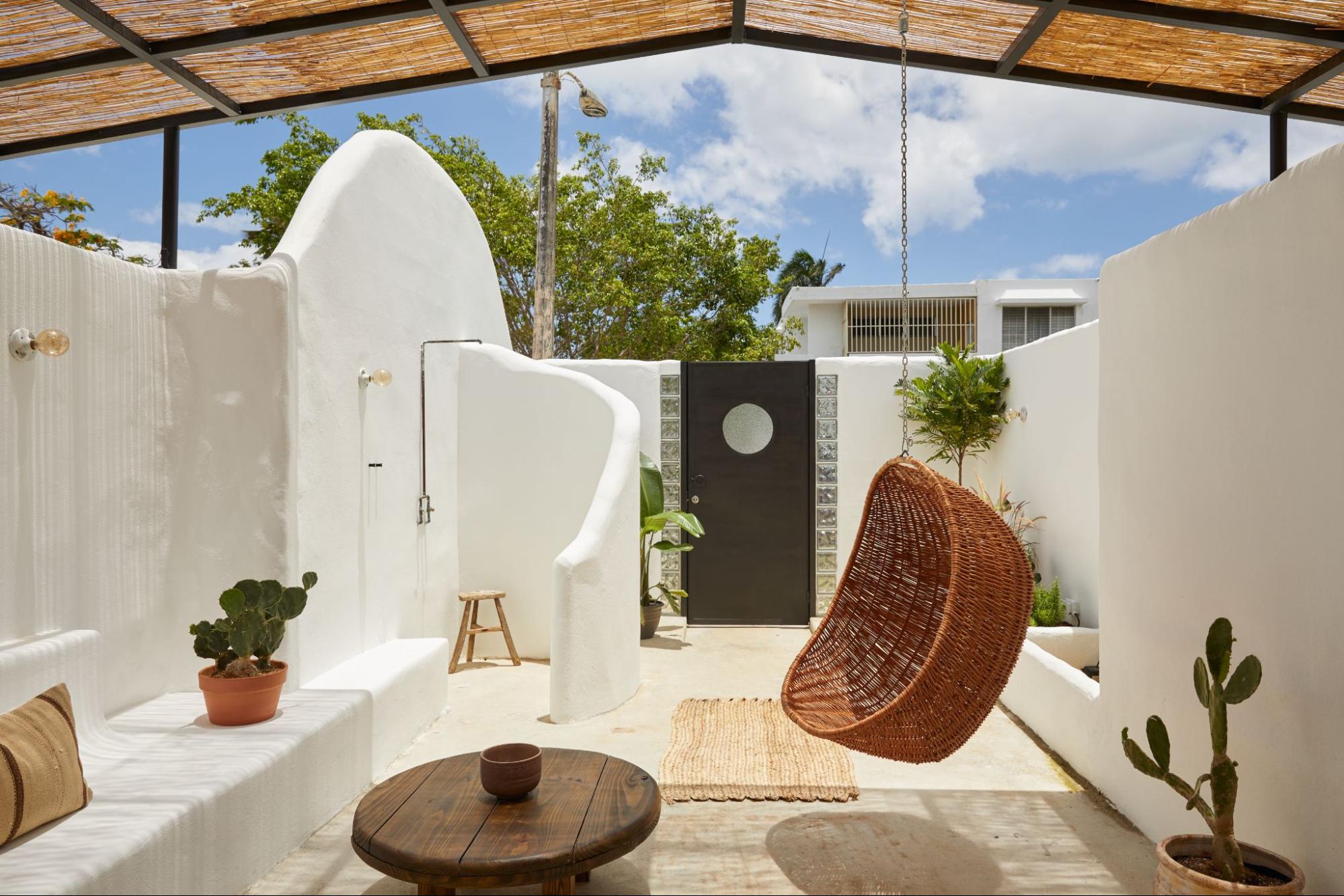 An open-air whitewashed patio courtyard featuring a hanging wicker chair, an outdoor shower, and a shaded pergola roof.