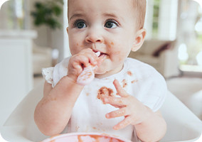 A child with food stains on his face and clothes sits next to the table, holding a spoon in his hands