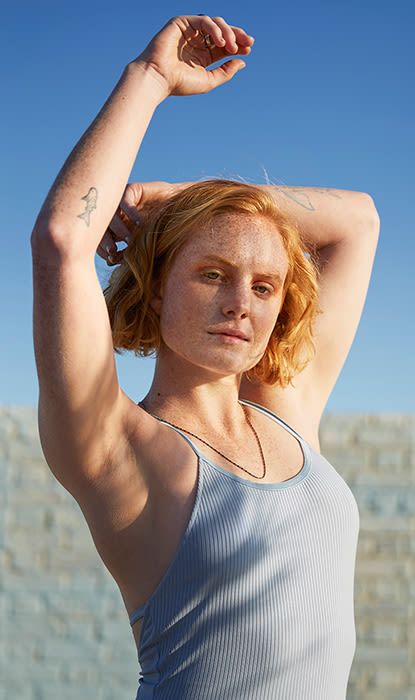 Una donna con i capelli corti e ondulati posa all'aperto, indossando una canotta azzurra sotto un cielo azzurro.