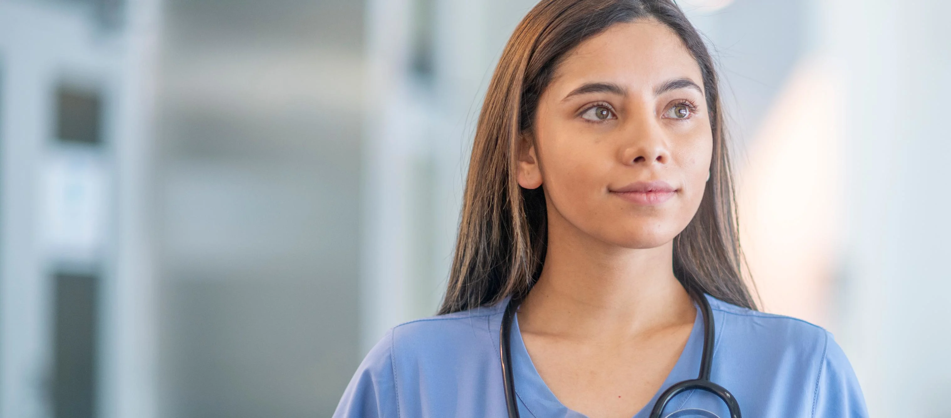 A young female medical student is standing inside a medical building. She is wearing blue medical scrubs and is looking away from the camera. GettyImages-1309503232.jpg