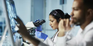 Photo of R&D researchers in a lab, one with a microscope and the other with a digital screen (Source: istock.com/poba)