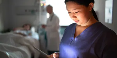 An Asian woman nurse checks patient charts on a digital tablet while her colleague tends to a patient in the background.