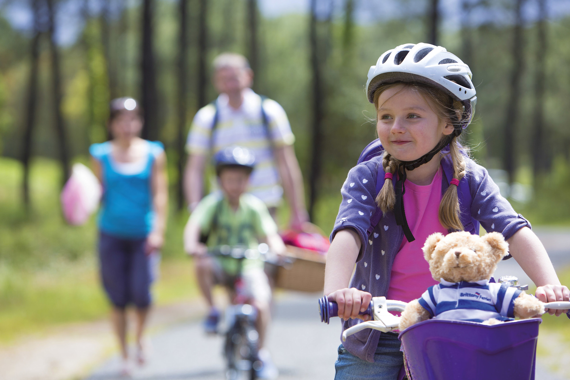 Girl on her bike with teddy bear ahead of her family on wooded track
