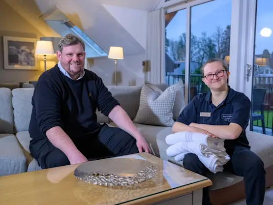 A man and woman sit next to each other on a sofa. The woman is holding some white bath towels.