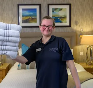 A young woman stands inside a hotel room and holds a pile of white bath towels in one hand.