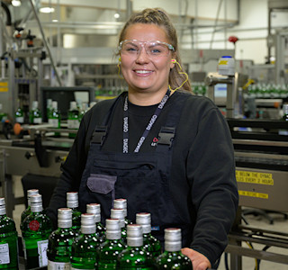 A woman, wearing safety goggles, next to a line of green bottles inside a factory.