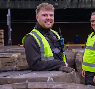 Two men who're wearing hi-vis vests stand next to whisky barrels.