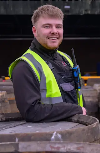 Two men who're wearing hi-vis vests stand next to whisky barrels.