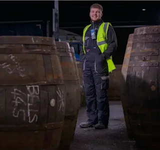 A young man stands next to some whisky barrels.