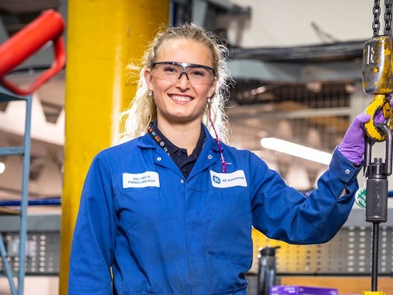 Young woman in blue overalls stands in front of machinery and smiles at the camera