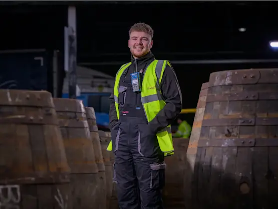 A young man wearing a hi-vis vest stands next to whisky barrels.