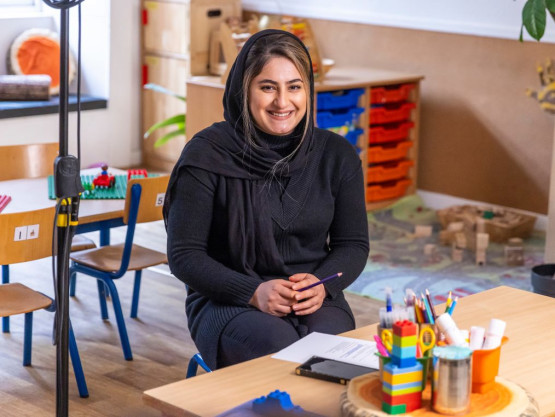 Young woman in black clothing sits at a table in a children's nursery and smiles to camera