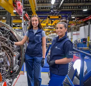 Two young women stand next to an aircraft part.