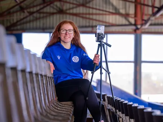 A young woman sits in the stand at Palmerston Park and holds a tripod with a smartphone attached.