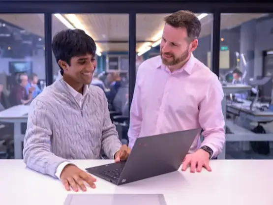 Two men stand at a desk and focus on a laptop screen.