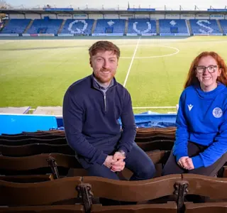 A man and a young woman sit in the stand at Palmerston Park. The football pitch is in the background.