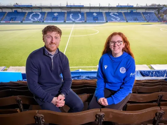A man and a young woman sit in the stand at Palmerston Park. The football pitch is in the background.