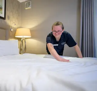 A young woman runs her hand along the top of a duvet within a hotel room.