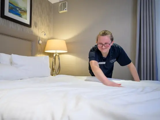 A young woman runs her hand along the top of a duvet within a hotel room.