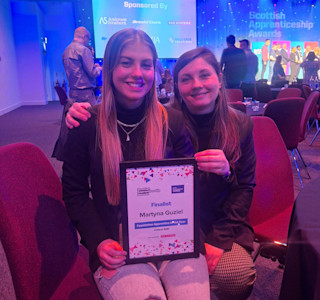 Two young women sitting down at a function. One woman holds a certificate that says 'finalist' on it.