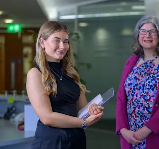 Two women stand next to each other in an office environment.