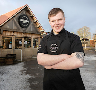Young smiling man in black work clothing. standing in front of farm shop