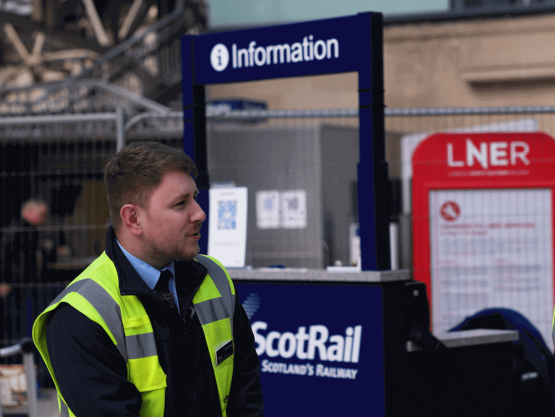 Two ScotRail workers chat in front of an information point in a train station.