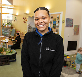A woman, wearing a black zipper, smiles at the camera while standing in a nursery environment.